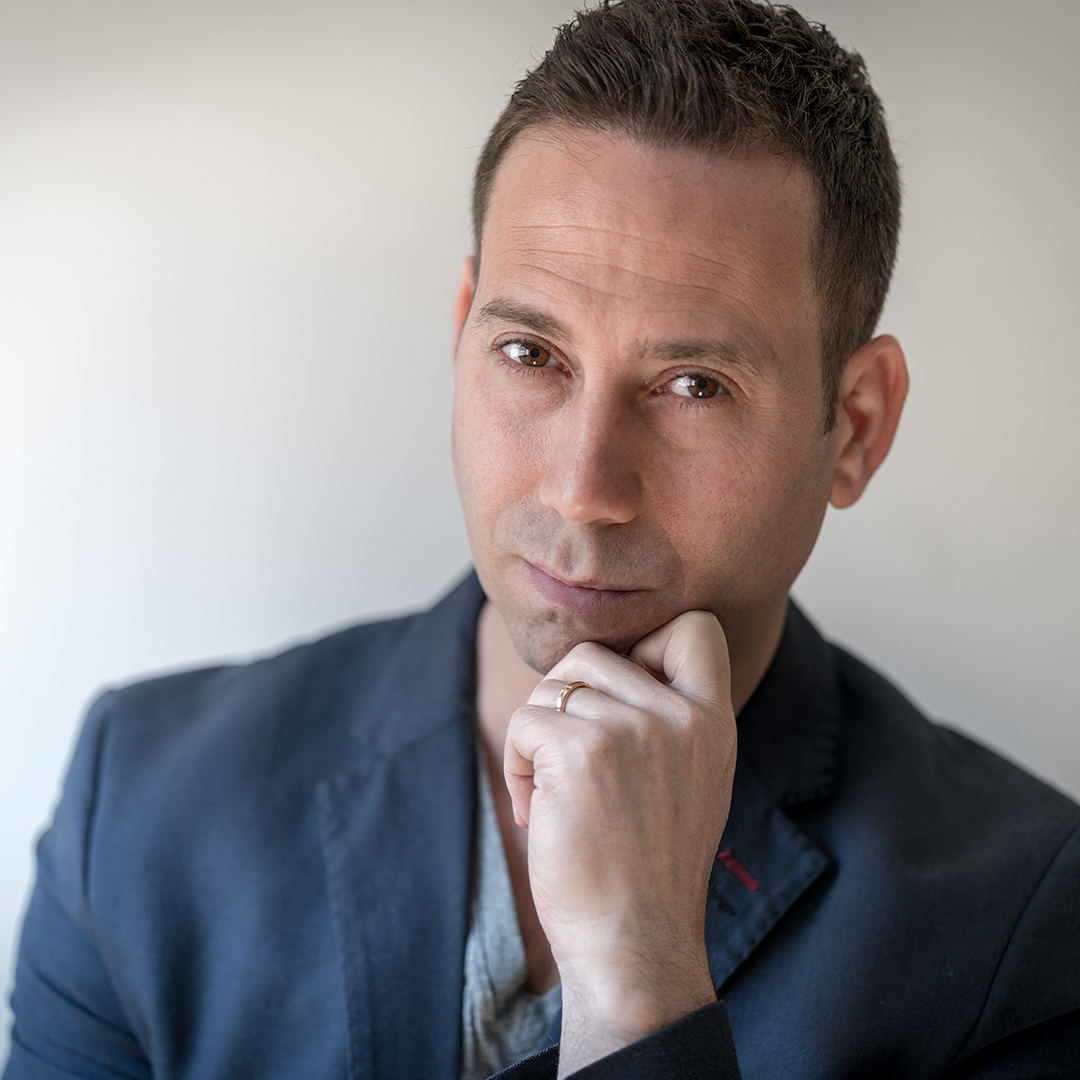 A man in a blue jacket posing with his hand on his chin.
