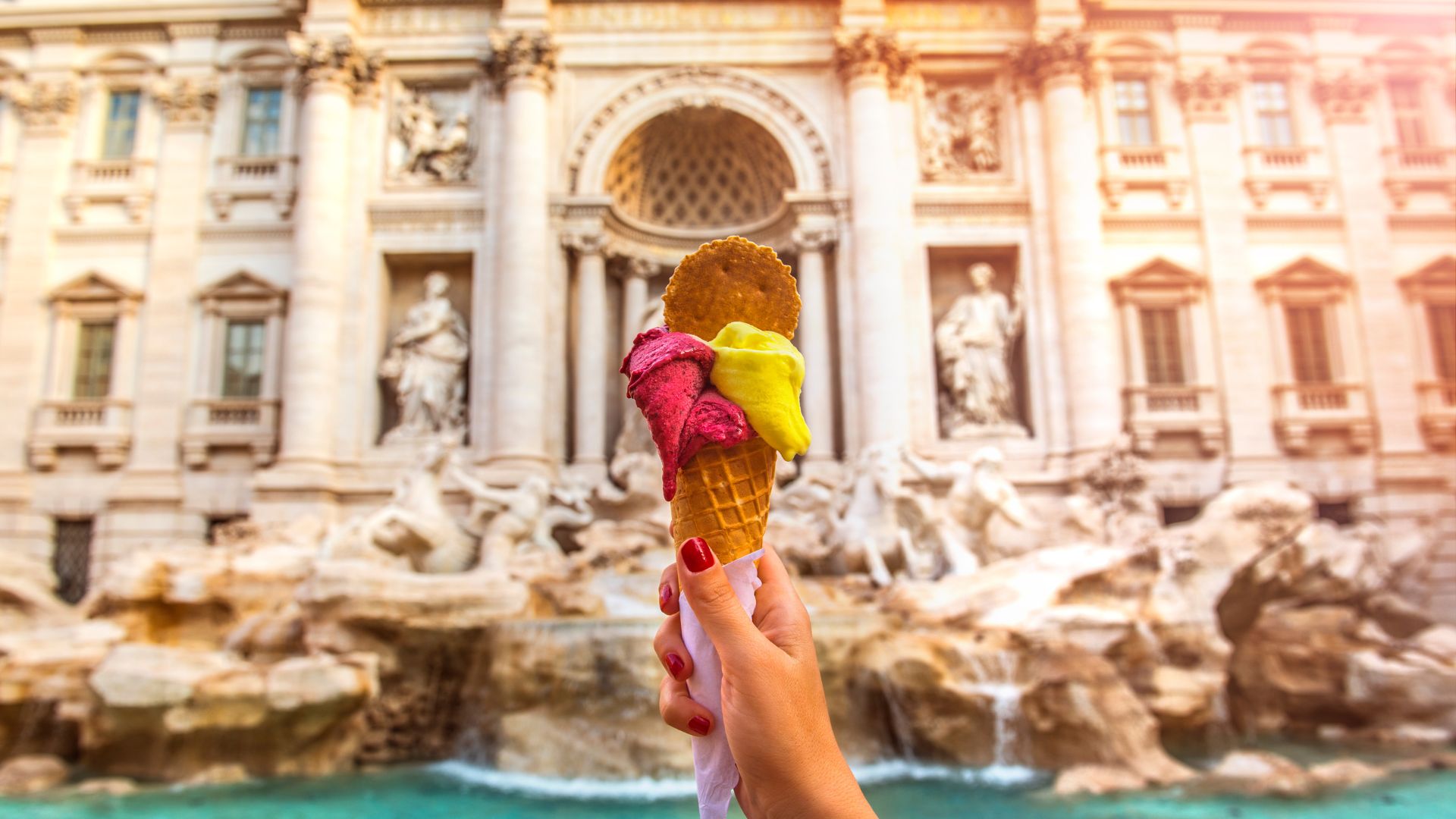 A hand holding an ice cream cone in front of Trevi fountain in Rome.