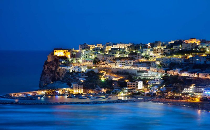 Night view of Peschici in Puglia, Italy, with illuminated buildings and the shimmering reflection of lights on the calm waters.